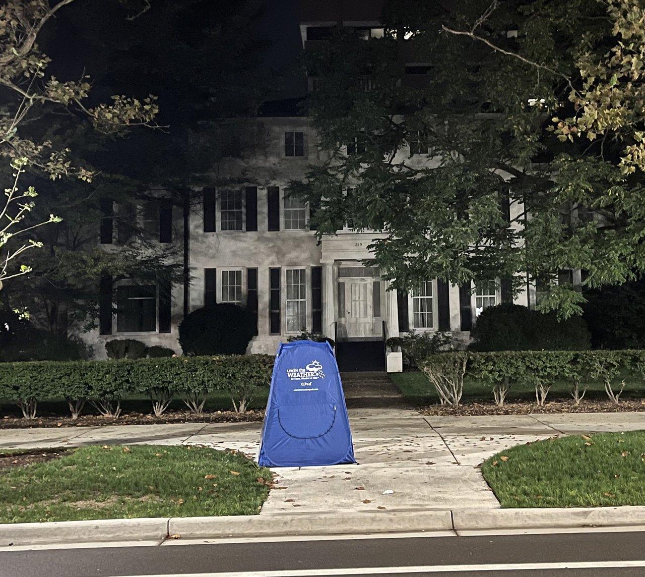 A small blue pop-up tent alone on the sidewalk at night, in front of the University of Michigan president's residence.